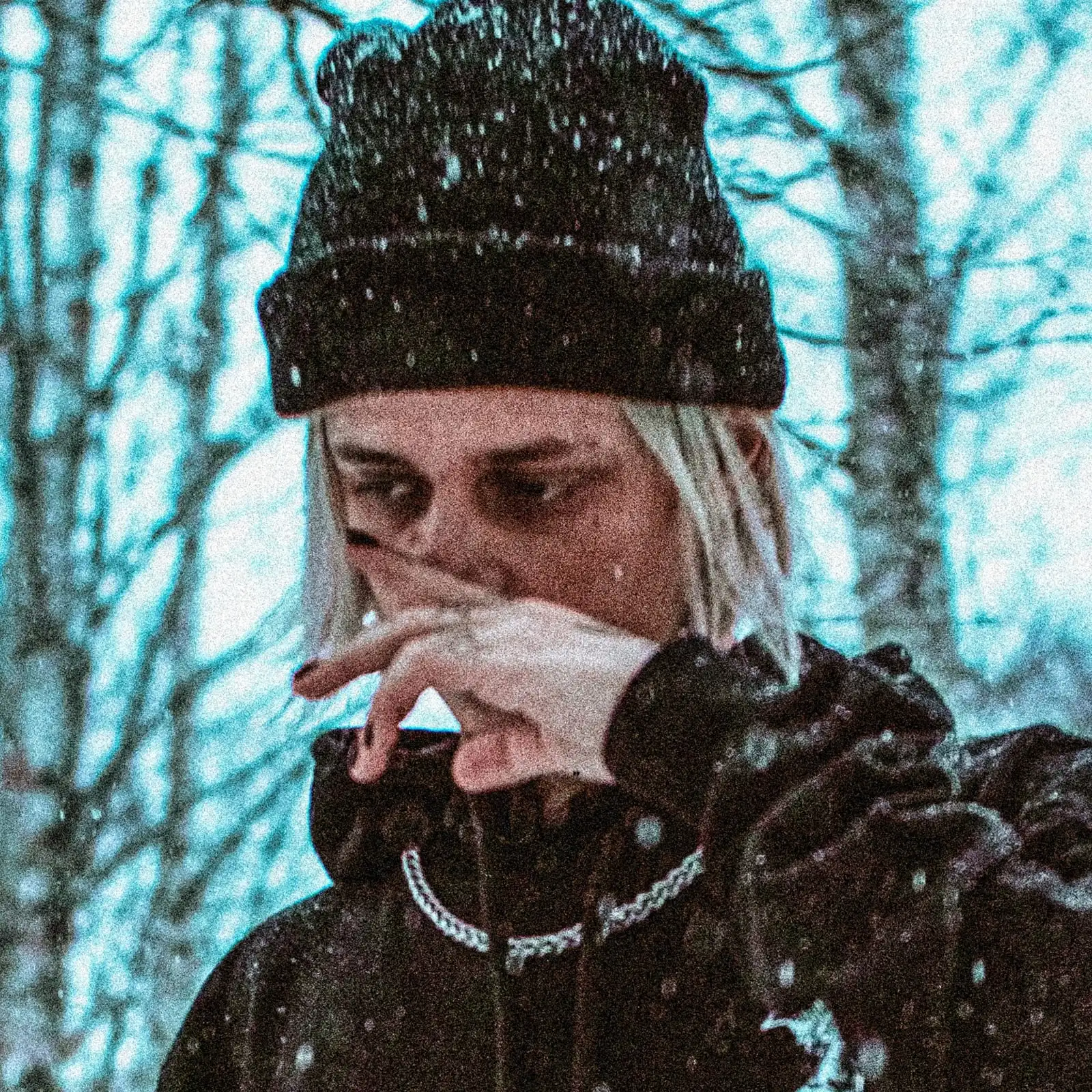 БЛЕСК in a black beanie, hand raised to the face, among snowy trees.